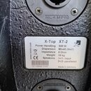 Detailní záběr na zadní část černého reproduktoru s texturovaným povrchem. Na bílém štítku je uveden model X-Top XT-2, značka s logem X a parametry:…