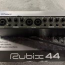 Černý USB audio interface s kovovým povrchem, umístěný na originální krabici. Na zařízení jsou viditelná loga Roland a nápis USB AUDIO INTERFACE Rubi…