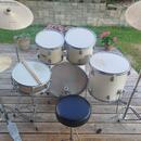 Celá bicí souprava v krémové barvě umístěná na dřevěné terase. Sestává z několika bubnů, dvou činelů značky MEINL LIVE SOUND a černého sedáku. Na jed…