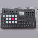 Černý digitální drum machine s ovládacími knoflíky, tlačítky a displejem. Na přístroji je v pravém horním rohu logo Elektron a na displeji nápis Anal…