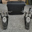 Dvojpedál na bicí buben v černé barvě s kovovými mechanismy a plastovým kufrem. Na obou šlapkách je bílý nápis IRON COBRA a pod ním kurzivou The Lege…