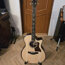 Akustická kytara s cutawayem, světlým topem a tmavými boky a zadní stranou. Na hlavě hmatníku je bílý nápis Taylor. Nástroj je umístěn ve svislé polo…