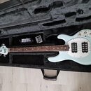 Světle modrá elektrická basová kytara v černém polotvrdém kufru. Na hlavě nástroje je bílý nápis MUSIC MAN. Kytara má dva snímače s kovovými kryty a…