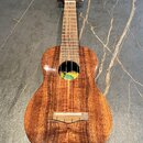 Dřevěný čtyřstrunný nástroj typu ukulele s hnědým povrchem a viditnou strukturou dřeva. Na hlavě je bílý nápis "Kala". Uvnitř ozvučovacího otvoru je…