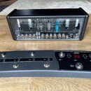 Černý kytarový zesilovač s průhledným předním panelem, za kterým jsou vidět elektronické součástky. Na skle v horní části je bílé logo Hughes & Kettn…