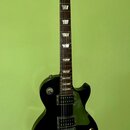 Černá elektrická kytara s tmavým hmatníkem a trapezoidními vložkami. Na hlavě je zlatý nápis Gibson a pod ním podpis Les Paul. Nástroj má dva humbuck…