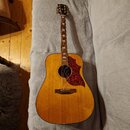 Akustická kytara s přirozenou dřevěnou barvou a hnědým hmatníkem. Na hlavě je viditelné logo Epiphone. Kytara má červený zvířecí pickguard s texturou…