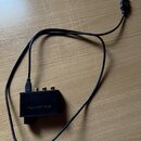 Černý obdélníkový adaptér s připojeným USB kabelem. Na přední straně je bílý nápis nuFORCE a tři zlaté konektory. Povrch zařízení vykazuje mírné zneč…