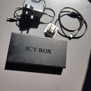 Černý obdélníkový box s nápisem ICY BOX. K zařízení patří černý síťový adaptér, bílý redukční adaptér do zásuvky a černý USB kabel. Povrch boxu vykaz…