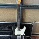Černá elektrická kytara typu Telecaster s bílým pickguardem a světle hnědým krčkem. Na hlavě je viditelné logo Fender. Kytara leží v otevřeném černém…