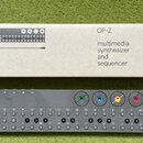 Šedý kompaktní syntezátor s nápisem OP-Z a čtyřmi barevnými ovládacími knoflíky (zelený, modrý, žlutý a červený). Zařízení obsahuje řadu malých kulat…