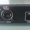 Černý kovový přístroj s nápisem MADI-USB v pravém horním rohu. Na předním panelu se nachází BNC konektor s nápisem MADI, USB port s označením USB a m…