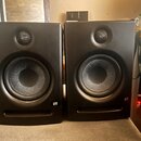 Dva černé studiové monitory v plastovém provedení. Na pravém dolním rohu každého reproduktoru je bílé logo "AMG". Povrch je matný s viditelnými prach…
