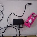 Růžový efekt pedál s nápisem DELAY®, ovládacím knoflíkem Time a přepínačem echo / f. back. Na pedálu je vidět logo ZFX a nápis DC 9V. Vedle pedálu je…
