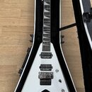 Bílá elektrická kytara s tvarem V v černém tvrdém pouzdře. Na hlavě hmatníku je čitelné logo Jackson. Kytara má dva humbuckery, Floyd Rose tremolo sy…