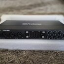 Černý audio rozhraní s modrým lemováním, umístěný v bílém polystyrenovém obalu. Na horní a přední straně je bílé logo PreSonus, vlevo nápis STUDIO 18…