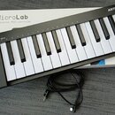 Černý kompaktní MIDI kontrolér s bílými a černými klávesami. Na levé straně je nápis MICROLAB a ovládací tlačítka s popisky Shift, Hold, Chord, Oct -…