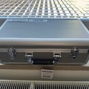 Stříbrný hliníkový kufřík s kovovými rohy, dvěma zámky a rukojetí. Na horní části je nalepené logo pedaltrain a čárový kód. Před kufříkem leží štítek…