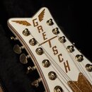 Detail hlavy bílé elektrické kytary s ozdobnými zlatými prvky. Na hlavě je svisle napsán nápis GRETSCH a jsou vidět zlaté ladičky. Povrch vykazuje dr…