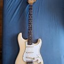 Elektrická kytara s krémovým trupem a bílým pickguardem. Na hlavě hmatníku je černý nápis Fender STRATOCASTER. Nástroj má tři snímače, tři ovládací k…