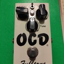 Bílý kytarový efekt s nápisy OCD® a Fulltone®. Na horní části jsou tři černé ovládací knoflíky s popisky Volume, Drive (částečně zakrytý) a Tone, dop…