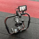 Na fotografii je černý stabilizátor typu gimbal s připevněným zrcadlovým fotoaparátem a externím monitorem. Na horní části ramene je viditelný červen…
