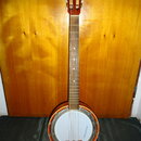Fotografie znázorňuje banjo s dřevěným hnedým tělem a hmatníkem z tmavého dřeva. Na bílé membráně je svisle napsaný modrý nápis Musima. Nástroj vykaz…