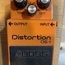 Kytarový efekt pedál oranžové barvy s černým spínacím pedalem. Na zařízení jsou nápisy Distortion, DS-1 a logo BOSS. Horní panel obsahuje tři ovládac…