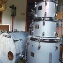 Sada bubnů v modrobílé barvě s chromovaným kováním. Na basovém bubnu je čitelné logo YAMAHA. Sestava obsahuje basový buben a tři tom-tomy naskládané…