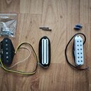 Fotografie zobrazuje tři elektrické snímače kytary v různých provedeních. Vlevo je černý snímač s viditelnými póly a připojeným žlutým a bílým kabele…