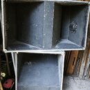 Dvě černé reproduktorové bedny s hliníkovým profilem na hranách. Povrch je z černého materiálu s viditelnými škrábanci, prachem a opotřebením. Na bed…