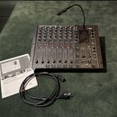 Černý mixážní pult s nápisem BEHRINGER PRO MIXER DX2000USB. Zařízení obsahuje několik kanálů s posuvníky, knoflíky a červenými tlačítky, v pravé část…