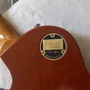 Detail zadní strany hřdelníku hnědé kytary z lakovaného dřeva. Na povrchu je圆形 černý štít s nápisy Gibson CUSTOM, EST. 1894 a MADE IN U.S.A. Povrch n…