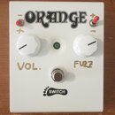 Bílý efekt pedál s nápisem ORANGE v horní části. Obsahuje dva bílé oválné ovládací knoflíky s popisky VOL. a FUZZ, uprostřed zelenou kontrolku a ve s…