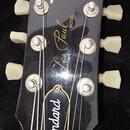 Detail hlavy černé elektrické kytary s bílými ladicími kolíky. Na povrchu je bílý nápis Gibson a zlaté nápisy Les Paul MODEL a Standard. Lak vykazuje…