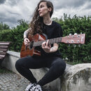 Žena drží v rukou akustickou kytaru s hnědým dřevěným povrchem a vyřezem v těle. Na hlavě kytary je bílý nápis „Blond“. Nástroj vypadá v dobrém stavu…