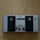 Stříbrný dvoupedálový spínač s nápisy A a B nad černými plasty pedálů. Uprostřed je logo BOSS, text DUAL FOOT SWITCH a model FS-6. V pravém horním ro…