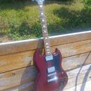 Tmavě červená elektrická kytara s tmelejším hmatníkem a bílými vložkami. Na hlavě je vidět nápis Gibson a logo značky. Kytara má dva humbuckery, stří…