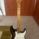 Elektrická kytara s tmavě hnědým průhledným lakem těla a bílým pickguardem. Na hlavě hmatníku je viditelné logo Fender. Hmatník a hlava jsou vyrobeny…