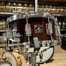 Tmavě hnědý dřevěný snare drum s chromovanými obručemi a mechanismy. Na korpusu je umístěn čtvercový štítek s nápisy TAMA, STARCASTING a MADE IN JAPA…