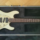 Elektrická kytara krémové barvy s perleťovým pickguardem, umístěná v černém tvrdém kufru. Na hlavě hmatníku je nápis MUSIC MAN a podpis Ernie Ball. N…