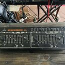 Černý elektronický syntetizátor s nápisy behringer a SYNCUSSION SY-1. Přední panel obsahuje posuvníky, otočné knoflíky a řadu tlačítek s popisky jako…