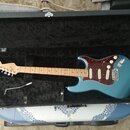 Modrá elektrická kytara typu Stratocaster s červeným trochlodňovým pickguardem v černém tvrdém kufru. Na hlavě kytary je nápis Fender STRATOCASTER. N…