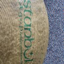 Detailní záběr na povrch mosazného činele s viditelnými rýhovanými drážkami. Na povrchu je zelený nápis „Istanbul Agop Handmade cymbals from Turkey“…