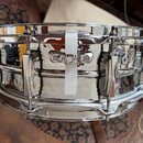 Chromovaný snare drum s kovovým korpusem azrcadlovým povrchem. Na přední straně jsou vidět chromované lugy a napínací šrouby. Uprostřed je umístěn bí…
