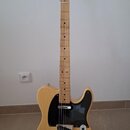 Světle žlutá elektrická kytara s černým pickguardem a javorovým hmatníkem. Na hlavě je viditelné logo Fender. Nástroj je umístěn na černém stojanu v…