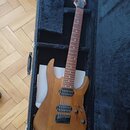 Elektrická kytara s přírodním dřevěným povrchem v černém tvrdém kufru. Na hlavě hmatníku je bílý nápis Ibanez. Nástroj má dva humbuckery, jeden ovlád…