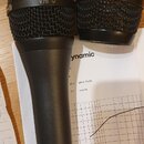 Dva černé dynamické mikrofony s mřížkou, jeden kompletní a jedna samostatná hlava. Na horním okraji mřížky prvního mikrofonu je nápis V70 a na mřížce…