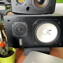 Černý studiový monitor s dřevěným povrchem. Na přední straně je viditelný bílý membránový basový reproduktor a menší výšek s černou mřížkou. V levém…