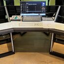 Bílý studiový stůl s černými doplňky a tmavě šedou polstrovanou hranou. Na stole je umístěn počítač s monitorem, bílá klávesnice, myš a stříbrný MIDI…